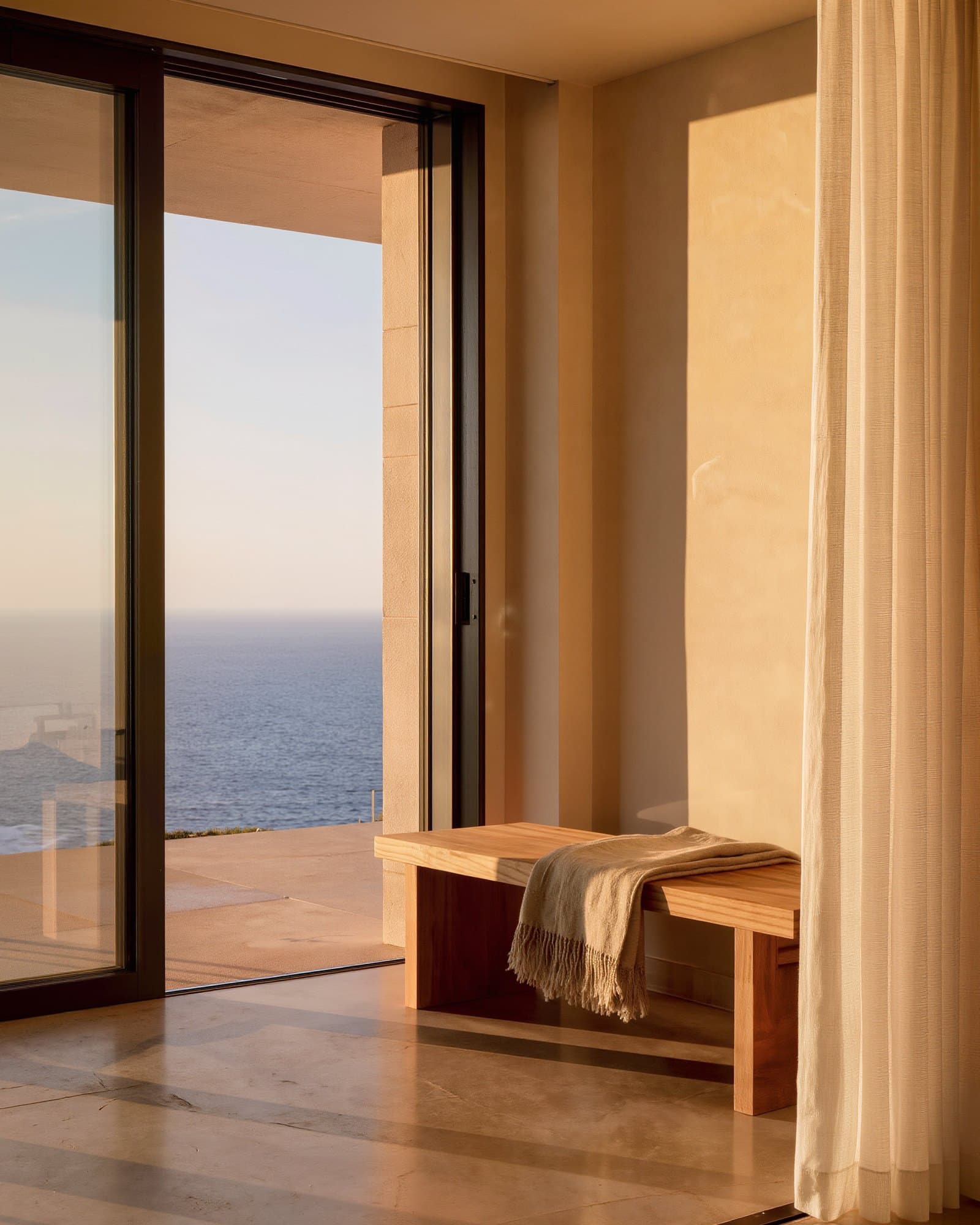 Wooden bench with a throw beside a floor-to-ceiling window onto the sea