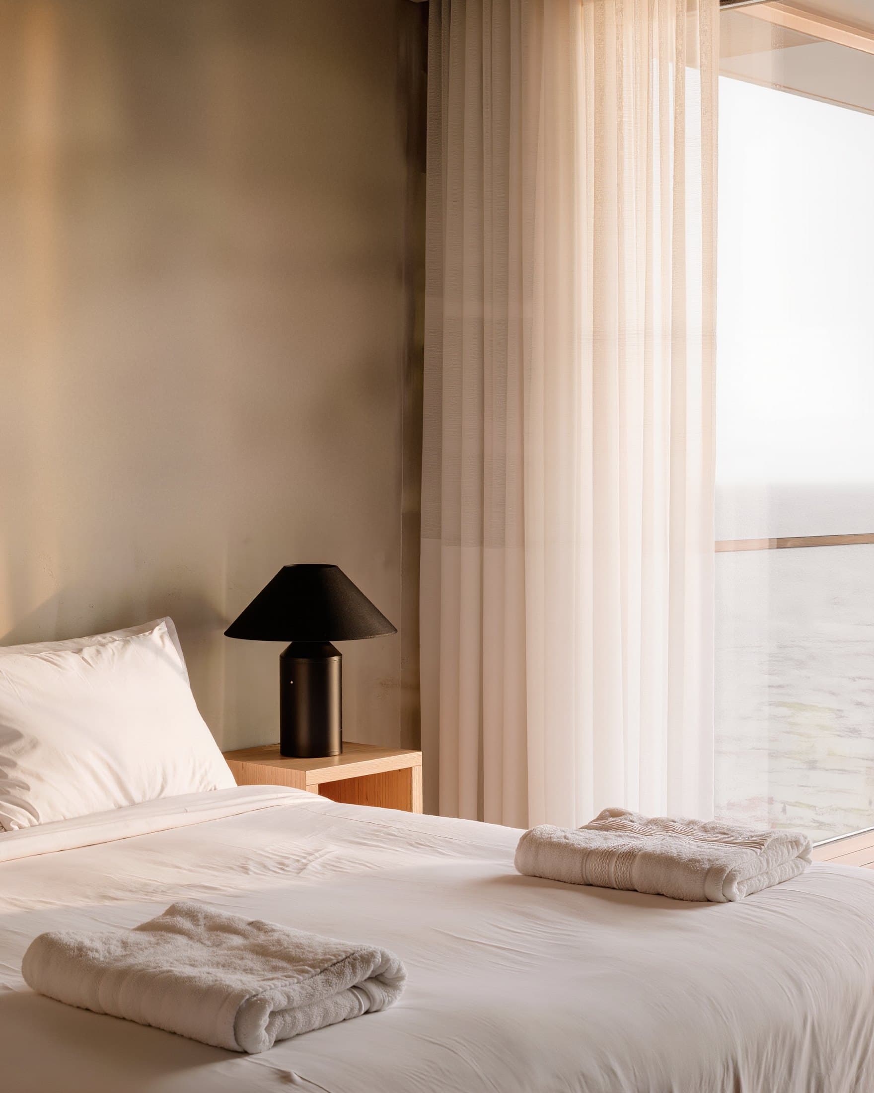 Cama recién hecha con toallas dobladas junto a un ventanal con vistas al mar